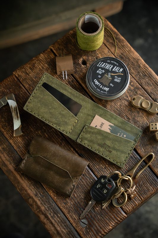 Olive leather bifold wallet with leather balm and keys on a workbench