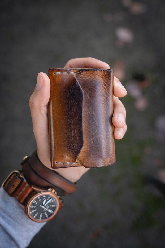 Hand holding a worn brown leather card wallet, bronze watch at the wrist