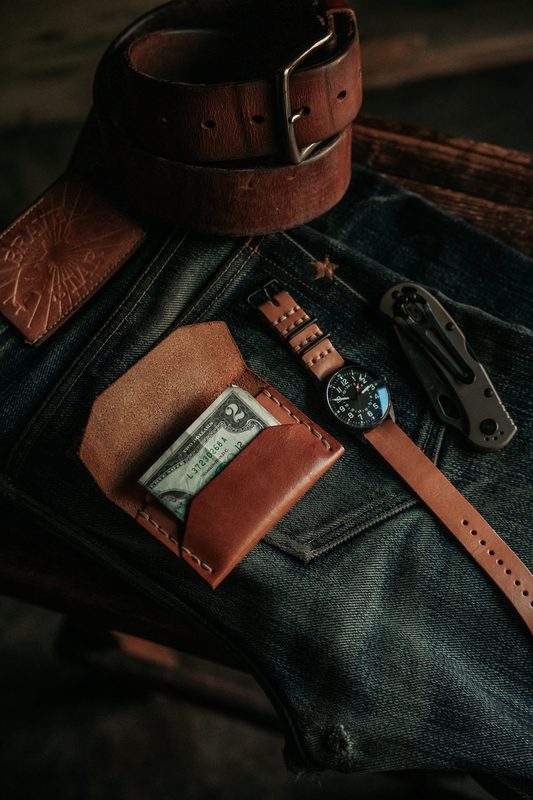 Leather card wallet with cash and a watch on denim, brown belt above