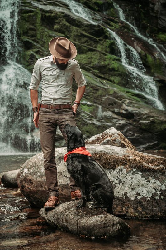 Man in a hat beside a waterfall with a black dog