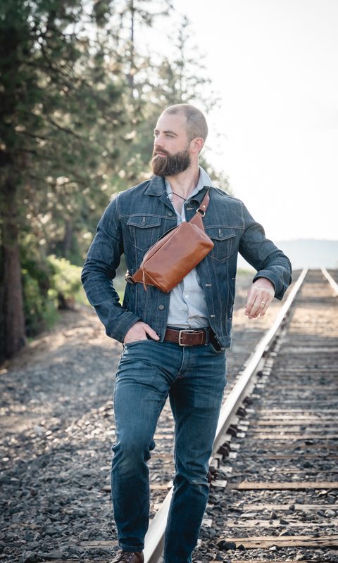Man in a denim jacket wearing a leather sling bag on railroad tracks