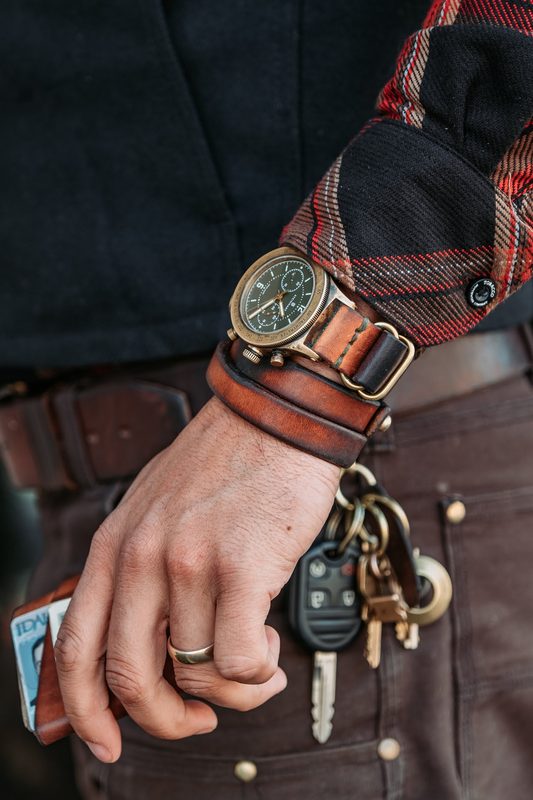 Bronze watch, leather wrist cuff and key clip against a brown work jacket