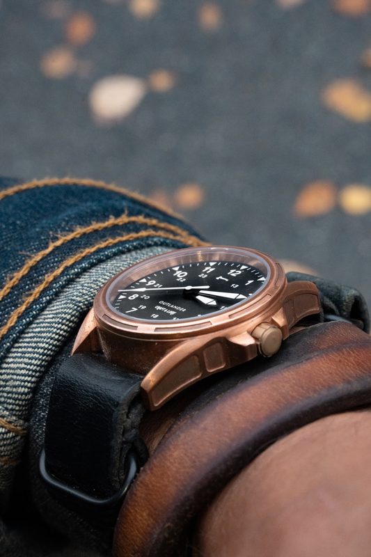 Close up of a bronze watch on a black strap with a leather cuff, autumn leaves below
