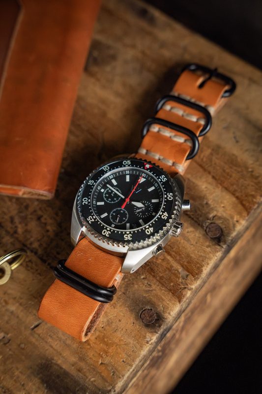 Black dial chronograph on a tan leather NATO strap on wood