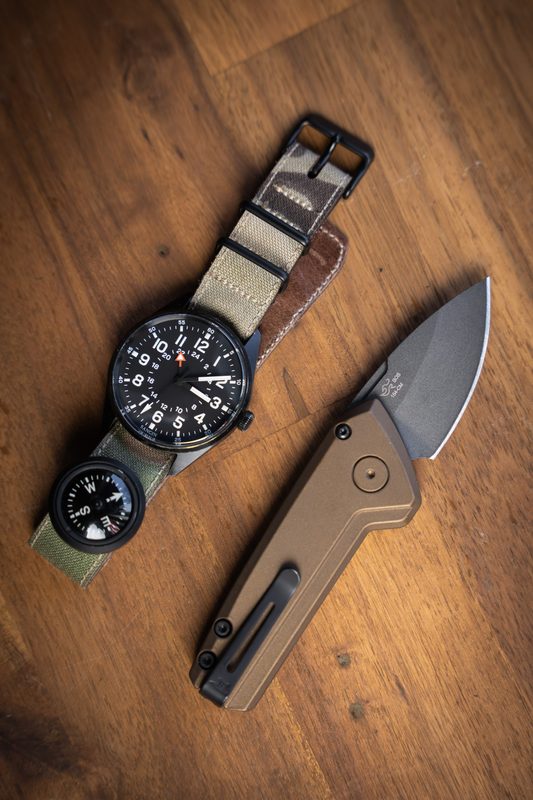 Chronograph on a camo strap beside a compass and folding knife on warm wood