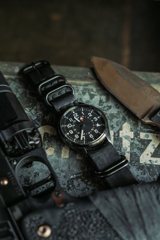 Black dial field watch on a black NATO strap beside a knife on a weathered crate
