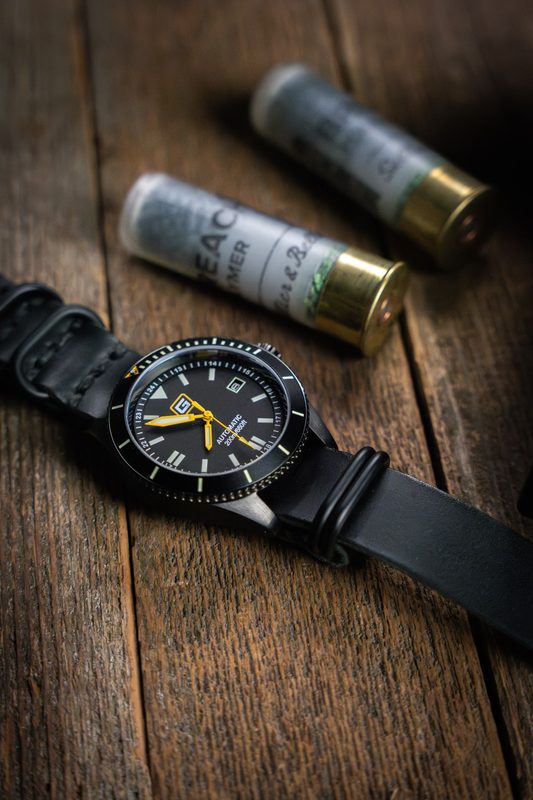 Black dive watch on a black strap beside shotgun shells on wood