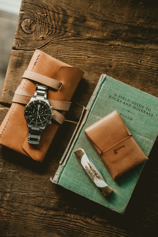 Steel watch on a leather watch pack with a card wallet and field guide on a wooden table