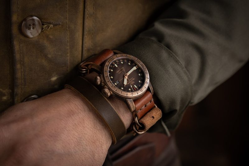 Bronze pilot watch on a brown leather cuff below a waxed canvas sleeve