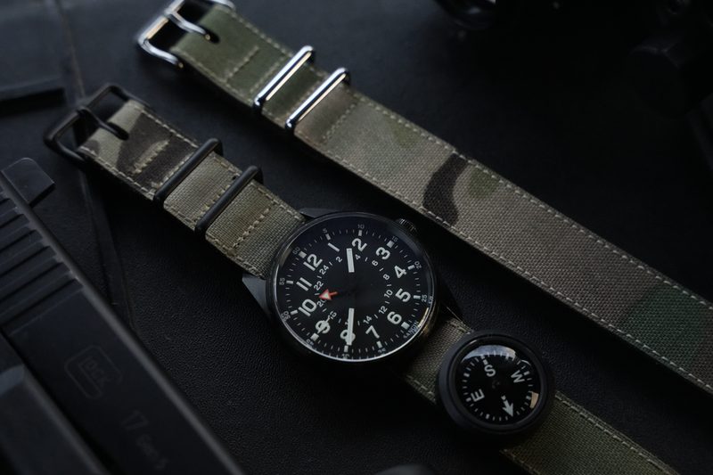 Black field watch on an olive canvas strap beside a compass on dark gear