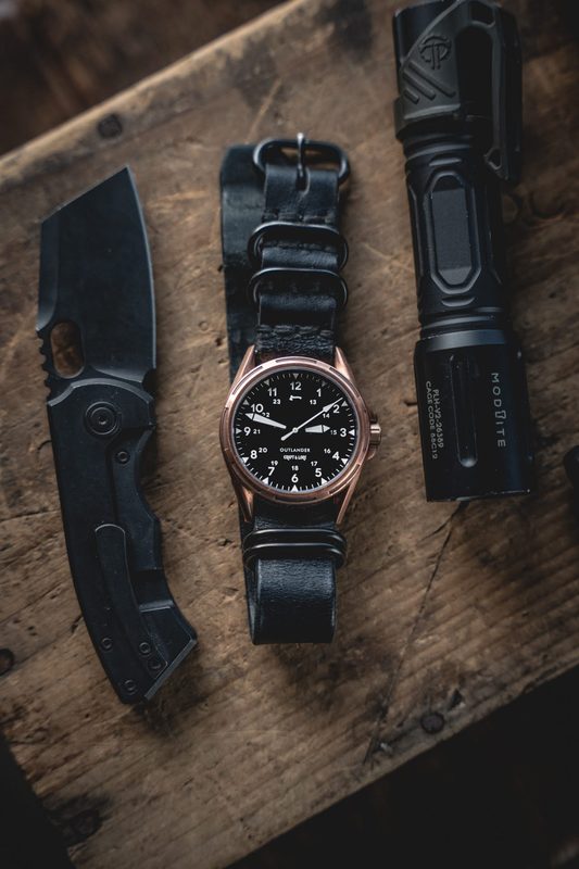 Bronze watch on a black NATO strap between a knife and flashlight on dark wood