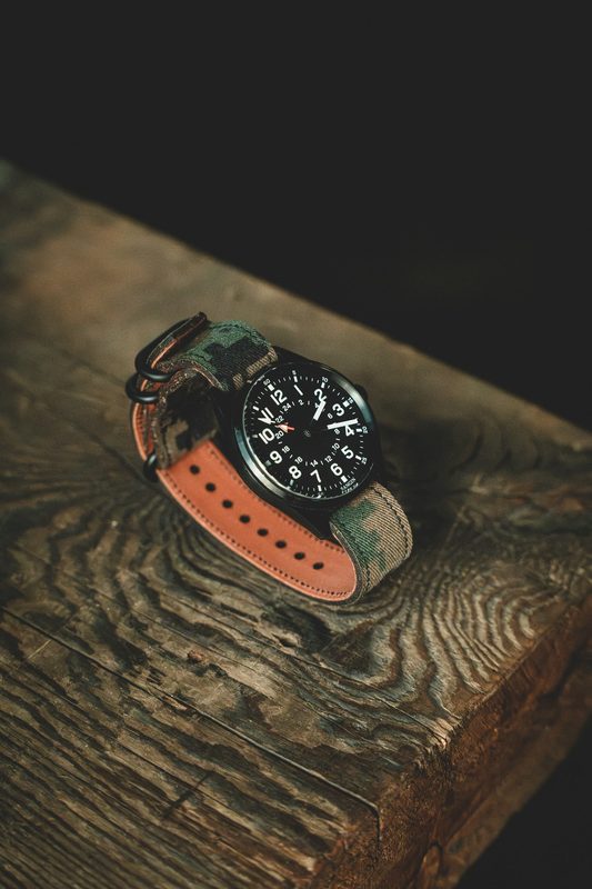 Black dial watch on a camo and orange leather hybrid strap on rough wood