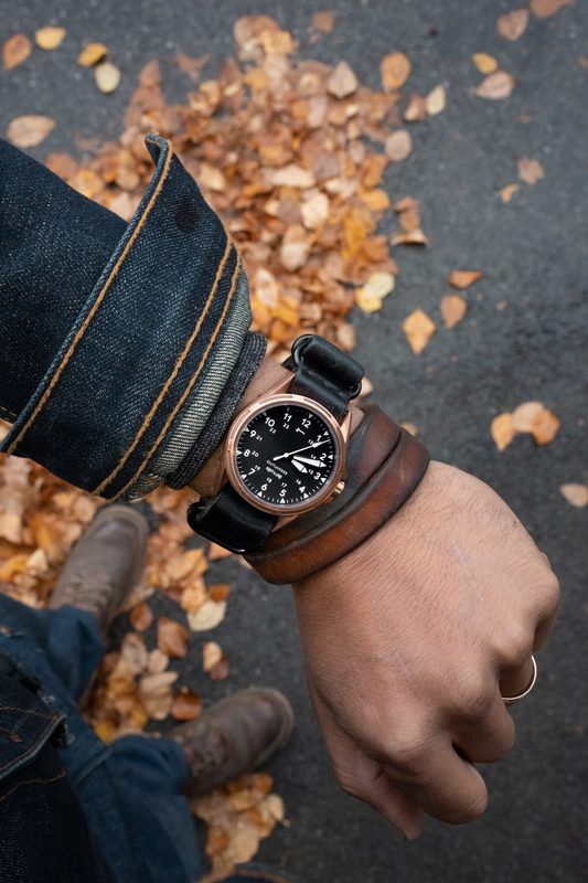 Bronze watch and leather cuff on the wrist over autumn leaves, raw denim sleeve