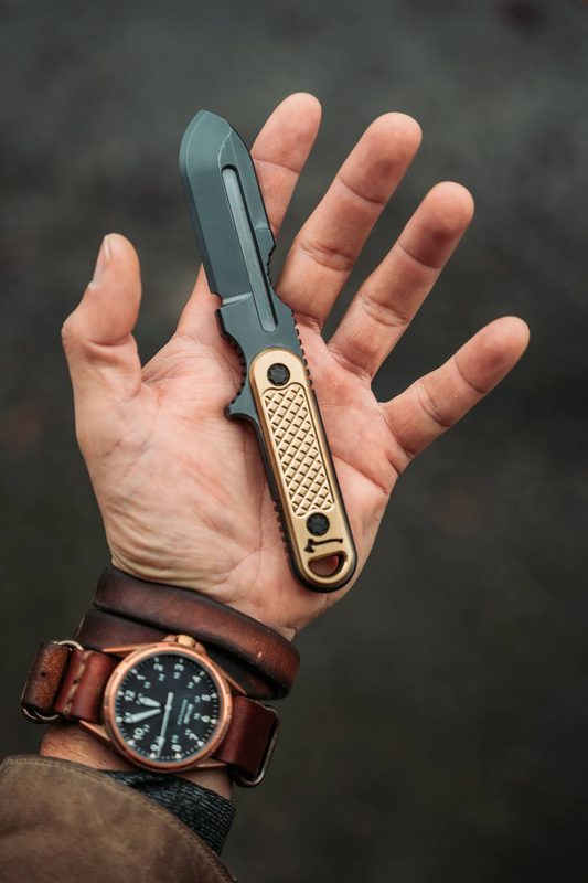 Hand holding a brass fixed blade knife, bronze watch on a leather cuff at the wrist