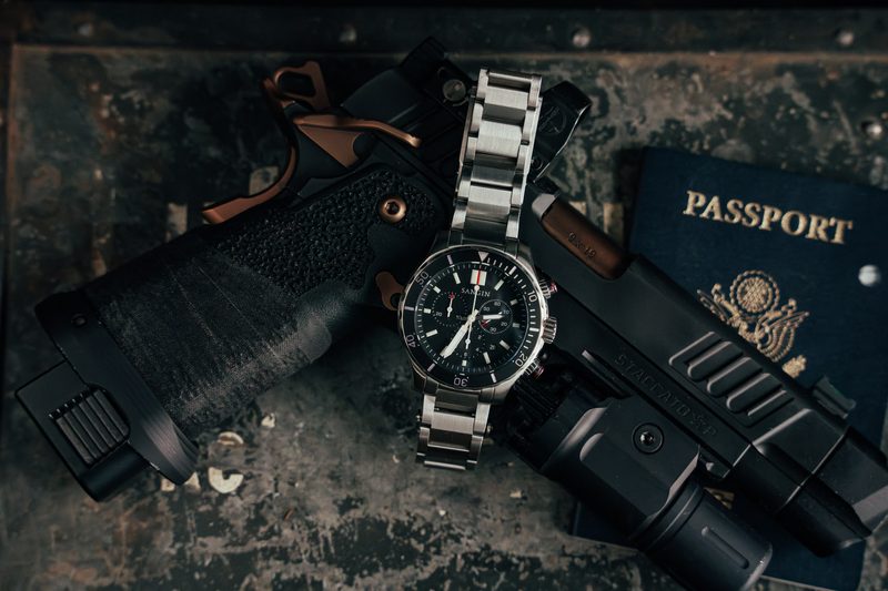 Steel chronograph on a bracelet beside a pistol and passport