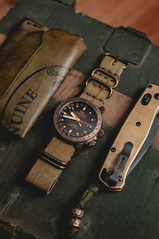 Flatlay of a bronze watch, leather sheath, brass comb, and folding knife on a green crate