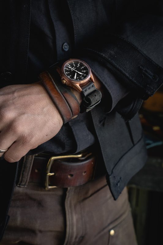 Bronze watch on a leather cuff at the wrist, hand resting on a leather belt