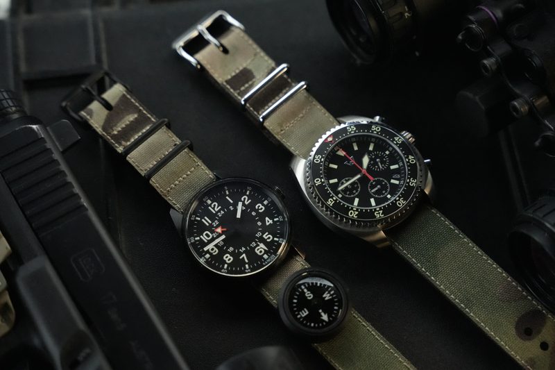 Two watches on olive canvas straps with a compass on dark gear