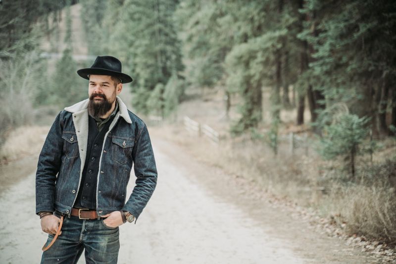 Chad in a denim sherpa jacket and black hat on a dirt road through pines